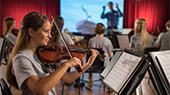 A teenage girl reads sheet music while practicing the violin in a youth orchestra setting