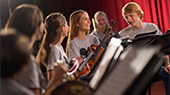 A group of teenagers with musical instruments smile while standing behind music stands in a studio setting